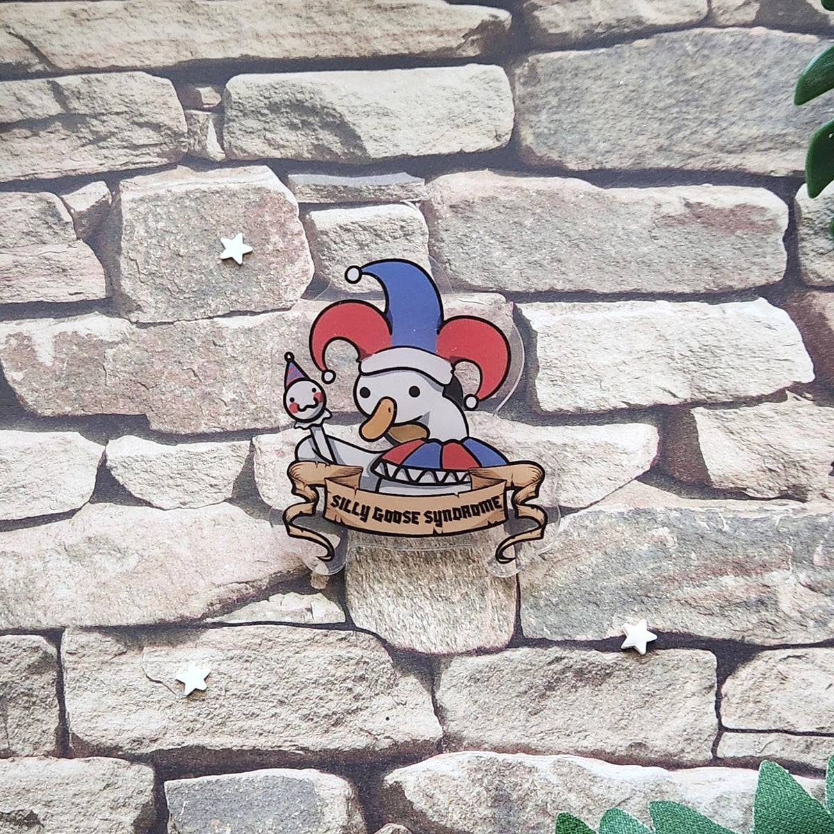 A pin featuring a cartoon goose in a jester hat with the text 'silly goose syndrome', laid on a stone backdrop.