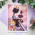 A printed anime art card featuring cat-themed characters on a cat tree, styled on a pastel purple background with green plant leaves around it.
