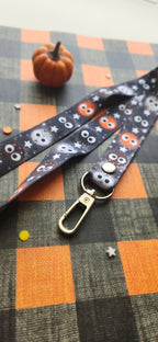 A Halloween-themed lanyard strap with a black background, printed with cute balls, and gray stars, featuring a gold metal lobster clasp. The strap is laid out on an orange and black plaid fabric surface next to a small decorative pumpkin.