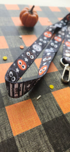 A Halloween-themed fabric lanyard strap with a black background, cute balls, and stars, laid diagonally on a black and orange plaid surface with a small pumpkin prop in the background.