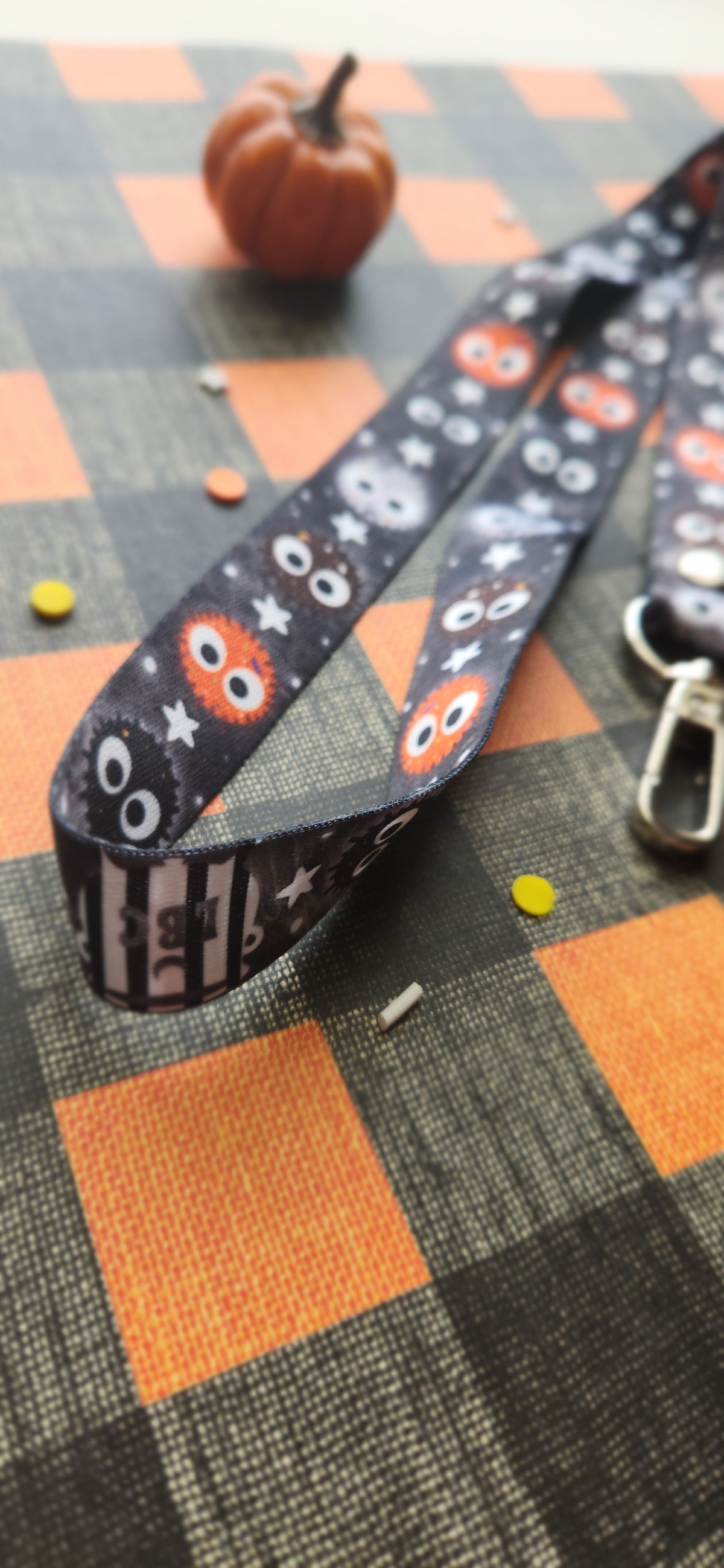A Halloween-themed fabric lanyard strap with a black background, cute balls, and stars, laid diagonally on a black and orange plaid surface with a small pumpkin prop in the background.