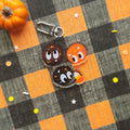 Three cute cartoon candy characters with large eyes and star details, attached to a silver lobster clasp, displayed on a black and orange buffalo plaid fabric with a small orange pumpkin prop.