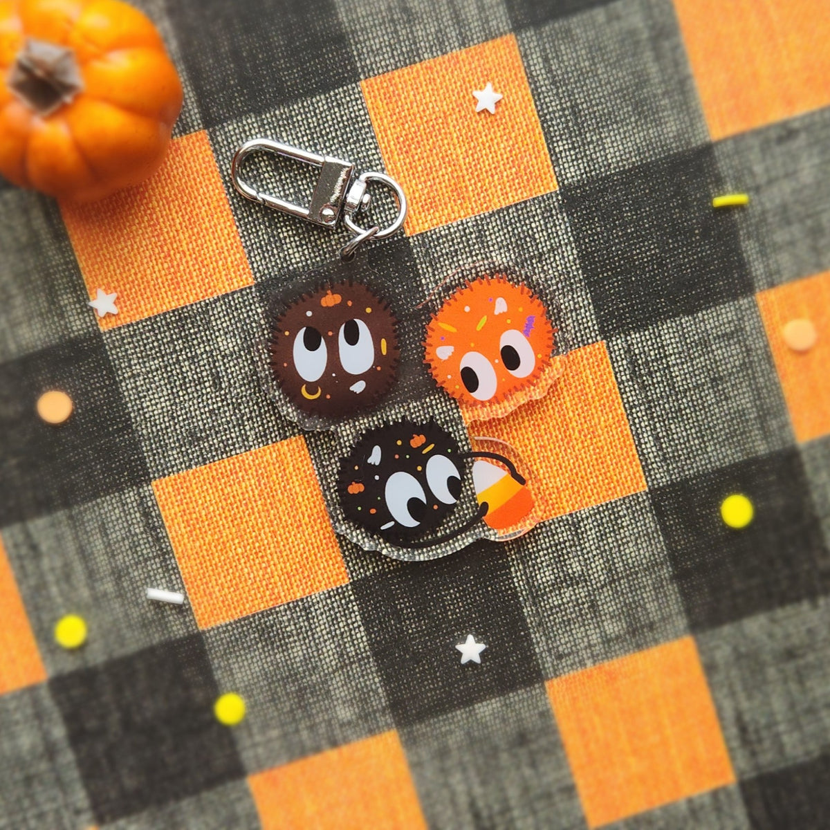 Three cute cartoon candy characters with large eyes and star details, attached to a silver lobster clasp, displayed on a black and orange buffalo plaid fabric with a small orange pumpkin prop.