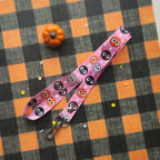 A pink fabric lanyard with a repeating cartoon ball pattern, laid flat on a black and orange checkered fabric background with a small orange pumpkin prop.