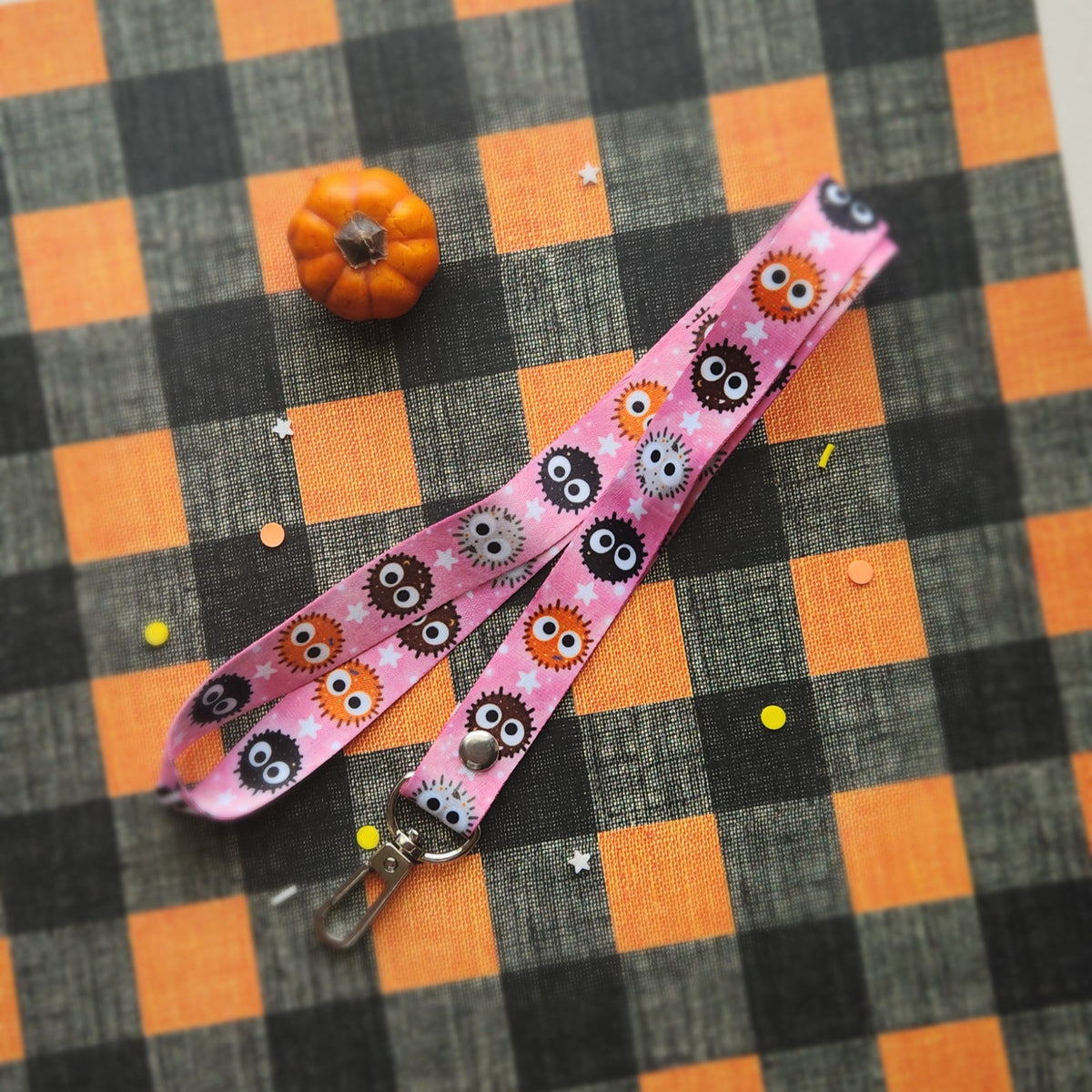 A pink fabric lanyard with a repeating cartoon ball pattern, laid flat on a black and orange checkered fabric background with a small orange pumpkin prop.