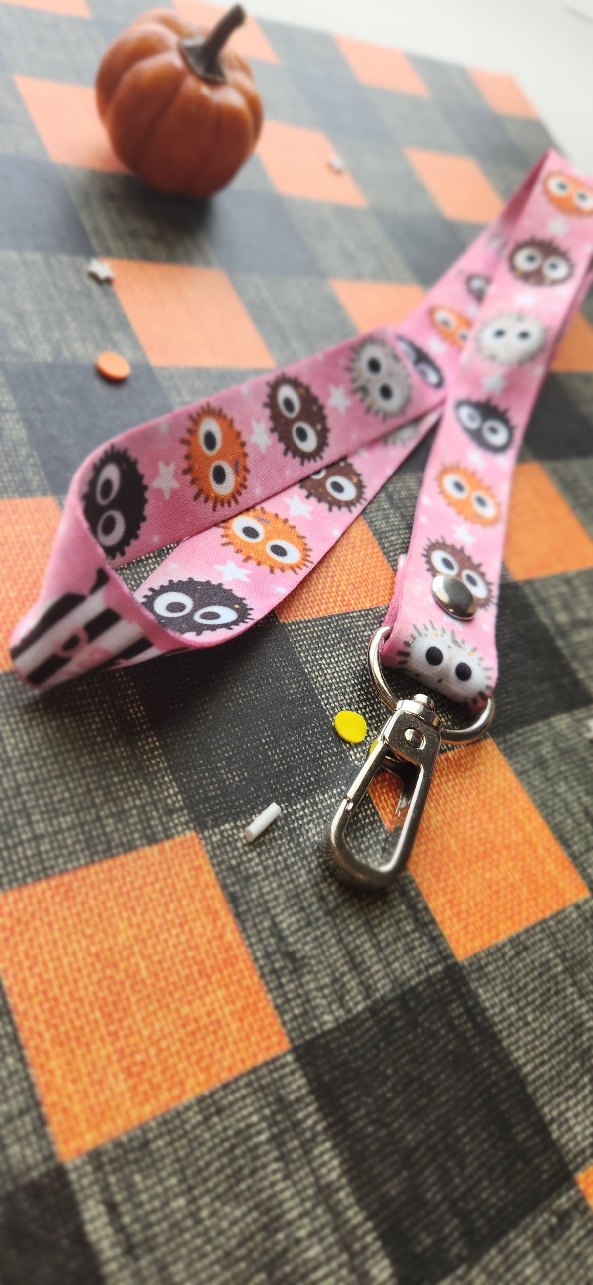 A pink fabric pet leash strap featuring a cartoon ball pattern, attached to a silver metal lobster clasp, displayed on an orange and black plaid blanket with a small pumpkin prop in the background.