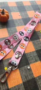 A pink fabric lanyard strap with a repeating pattern of cute cartoon balls, featuring a silver metal swivel snap and a silver lobster clasp, displayed on an orange and black buffalo plaid blanket.