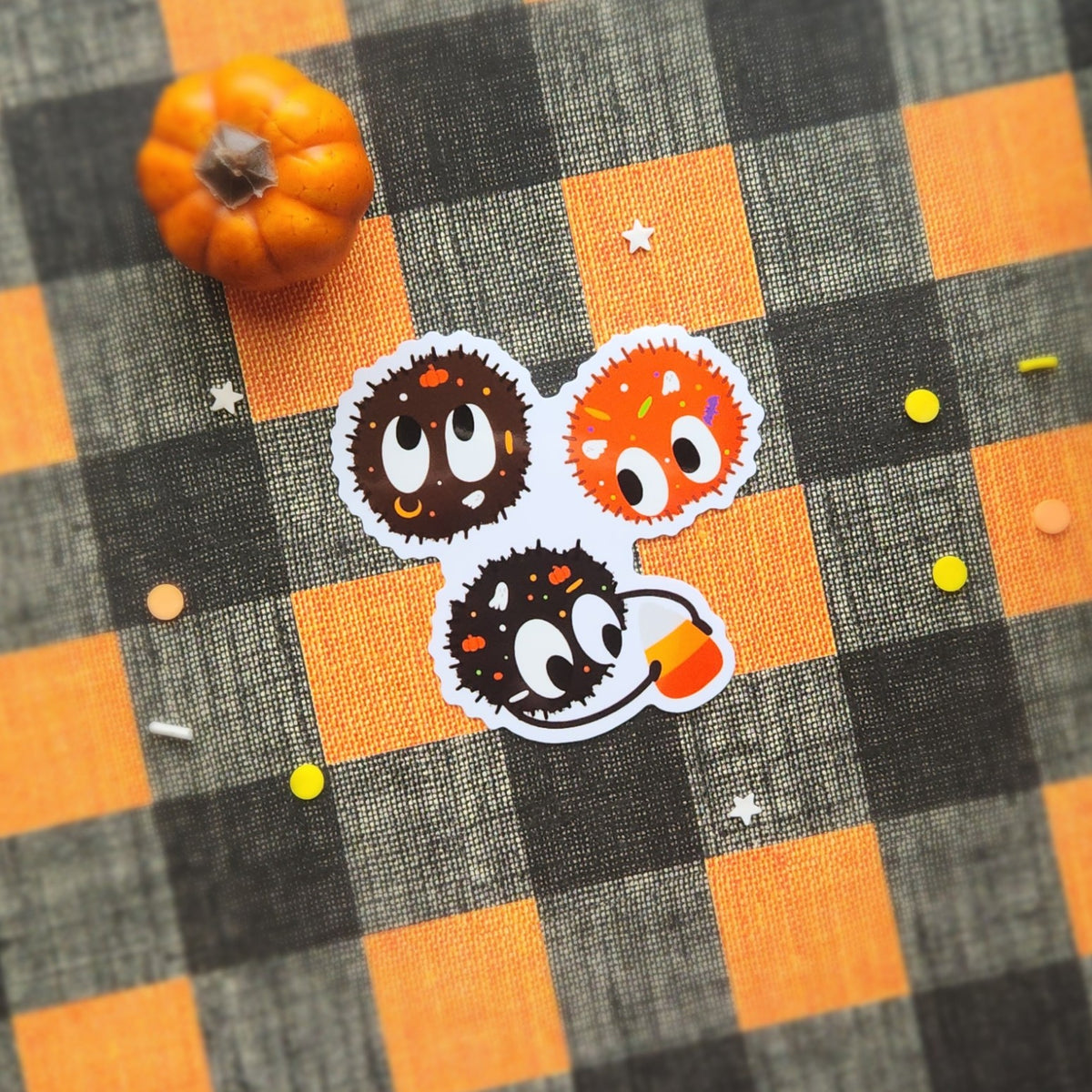 Three Halloween-themed character stickers arranged on a black and orange buffalo plaid fabric background, with a small orange pumpkin prop in the top left corner.