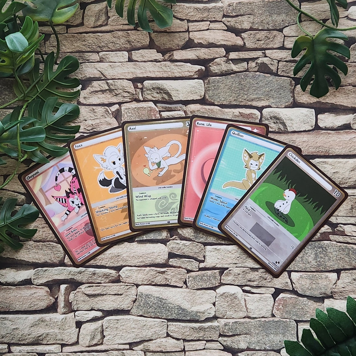 Five illustrated trading cards fanned out on a stone wall background with green leaves around the edges. The cards feature different fantasy animal designs and pastel-to-real color schemes.