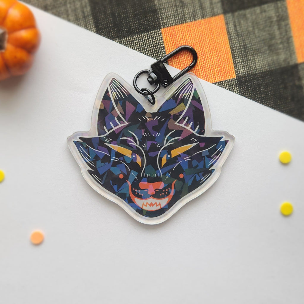 A colorful acrylic keychain featuring a stylized fox head in a geometric, mosaic-like pattern of blue, purple, black, and orange, attached to a black metal clasp. The keychain is photographed on a white surface with a black and orange plaid fabric, a small orange pumpkin, and small yellow and orange confetti in the background.