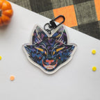 A colorful acrylic keychain featuring a stylized fox head in a geometric, mosaic-like pattern of blue, purple, black, and orange, attached to a black metal clasp. The keychain is photographed on a white surface with a black and orange plaid fabric, a small orange pumpkin, and small yellow and orange confetti in the background.