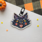 A die-cut acrylic keychain charm shaped like a stylized black cat head with a black metal clasp, photographed on a white surface with small orange and yellow confetti and a plaid fabric in the background.