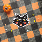A die-cut sticker featuring a stylized black cat head with yellow eyes and orange cheeks, placed on a black and orange buffalo plaid fabric background with a small orange pumpkin prop.