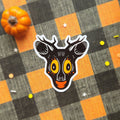 A die-cut sticker of a stylized deer head with antlers, featuring a black face, large yellow eyes, and an orange mouth, placed on a black and orange buffalo plaid fabric background with a small orange pumpkin prop.