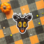 A die-cut sticker of a stylized deer head with antlers, featuring a black face, large yellow eyes, and an orange mouth, placed on a black and orange buffalo plaid fabric background with a small orange pumpkin prop.