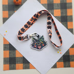 A Halloween-themed lanyard with a cat-shaped keychain, displayed on a white card over an orange and black buffalo plaid fabric background.
