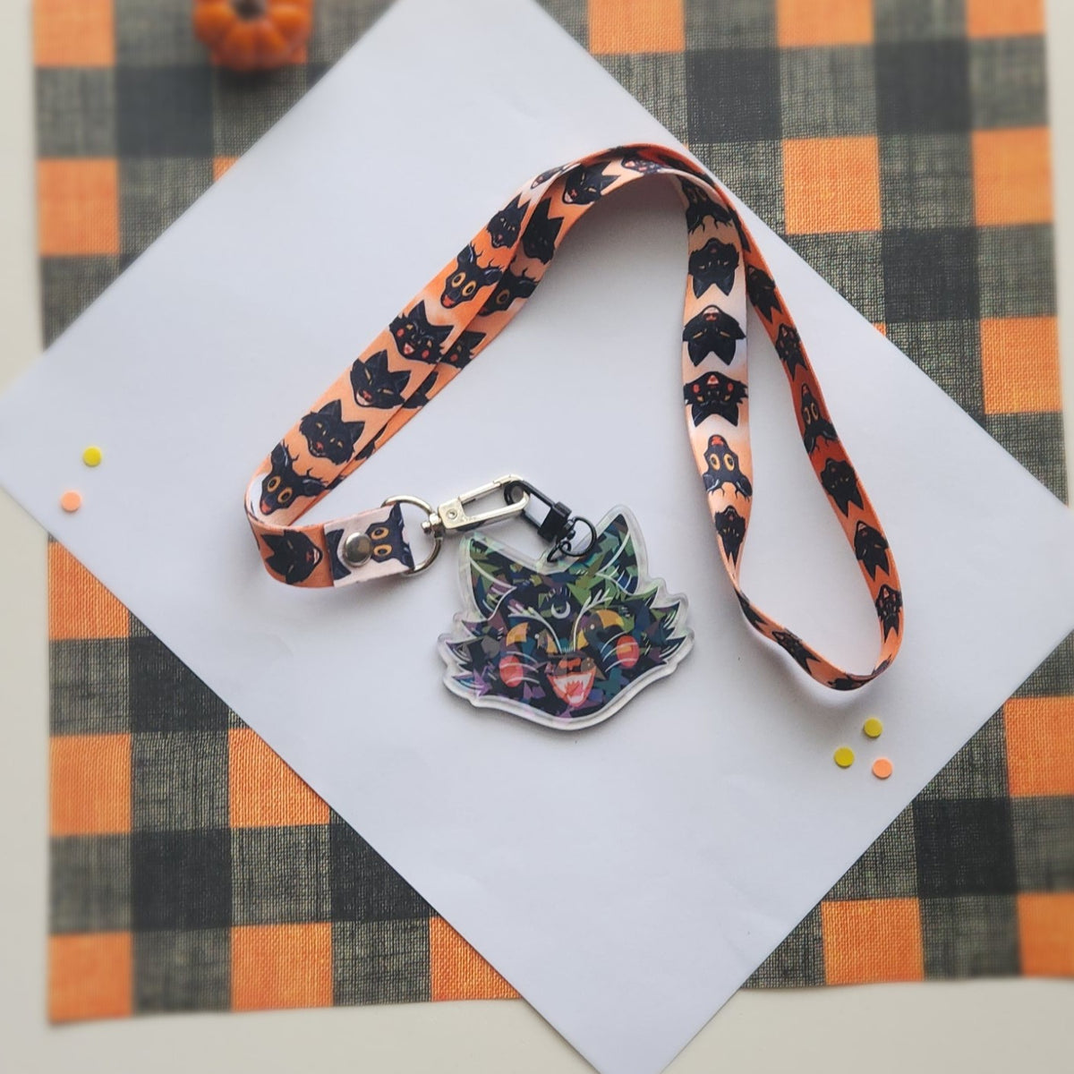 A Halloween-themed lanyard with a cat-shaped keychain, displayed on a white card over an orange and black buffalo plaid fabric background.
