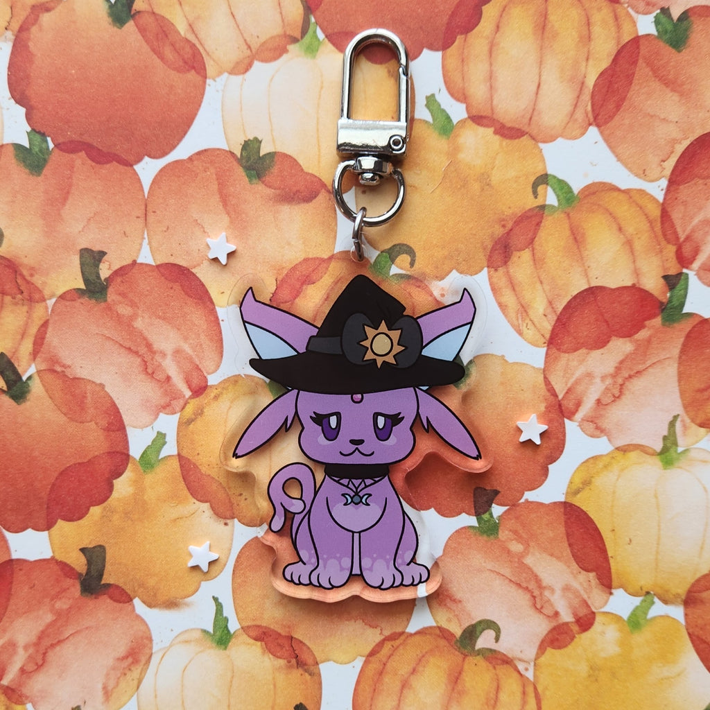 A purple cartoon fox charm wearing a black witch hat with a gold sun emblem, attached to a silver metal clasp, displayed on a watercolor pumpkin patterned background.
