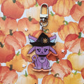 A purple cartoon fox charm wearing a black witch hat with a gold sun emblem, attached to a silver metal clasp, displayed on a watercolor pumpkin patterned background.