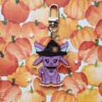 A purple cartoon fox charm wearing a black witch hat with a gold sun emblem, attached to a silver metal clasp, displayed on a watercolor pumpkin patterned background.