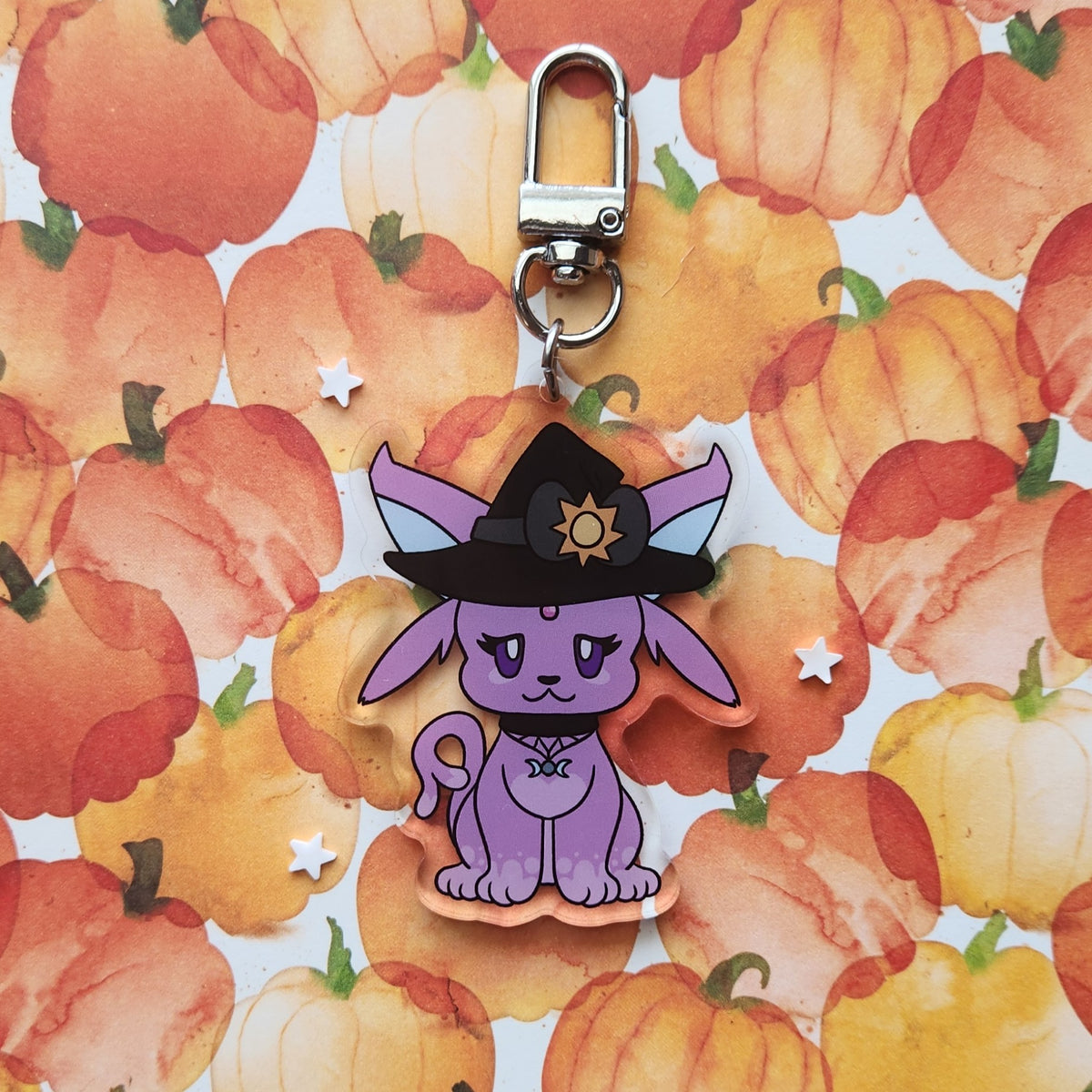 A purple cartoon fox charm wearing a black witch hat with a gold sun emblem, attached to a silver metal clasp, displayed on a watercolor pumpkin patterned background.
