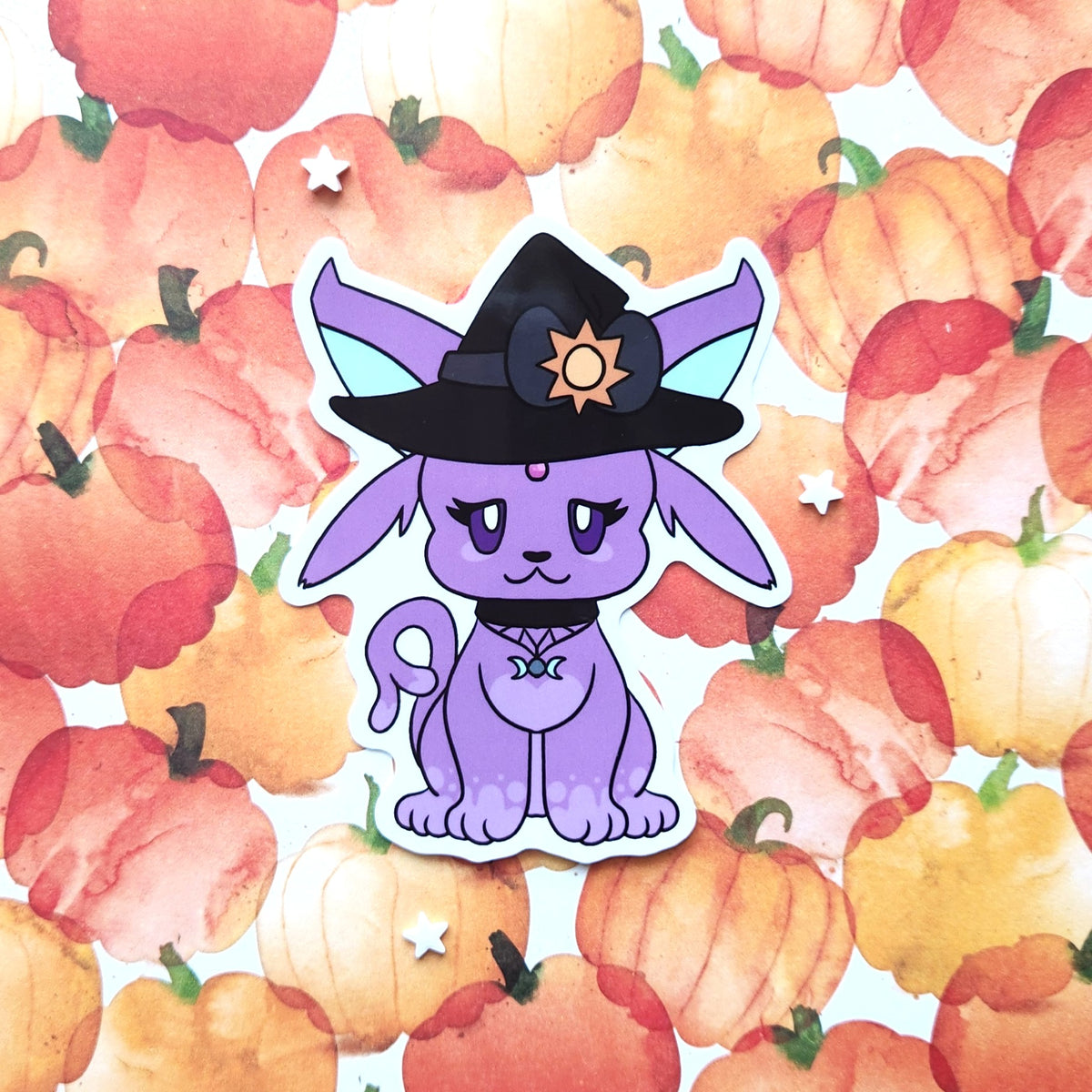 A die-cut sticker featuring a cute purple cartoon fox wearing a black witch hat with a gold sun emblem, set against a watercolor pumpkin pattern background.